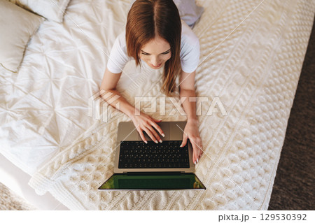 Young woman working on laptop while relaxing on cozy bed in a comfortable room during daytime Young woman working on laptop while relaxing on cozy bed in a comfortable room during daytime 129530392