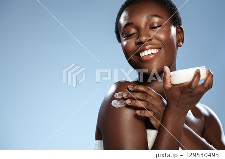Smiling woman applies cream on her shoulder against a clear blue background during a skincare routine 129530493