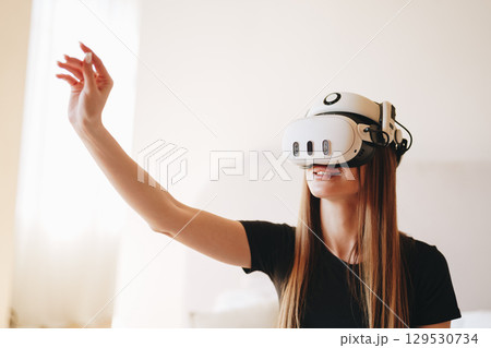 Young woman experiences virtual reality in a cozy indoor setting while smiling and interacting with digital elements 129530734