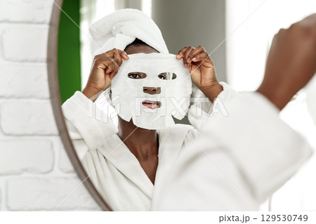 Woman applies sheet mask while looking into mirror in bright bathroom during self-care routine Woman applies sheet mask while looking into mirror in bright bathroom during self-care routine 129530749