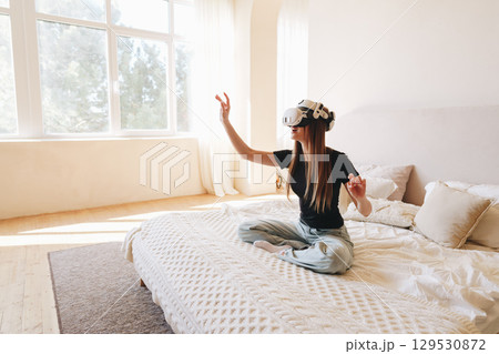 Young woman enjoys virtual reality experience while sitting on a bed in a bright room during daytime Young woman enjoys virtual reality experience while sitting on a bed in a bright room during daytime 129530872