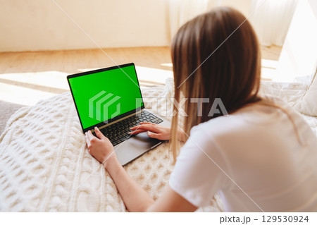 Woman sits on a cozy bed working on a laptop with a green screen during daylight in a bright room 129530924