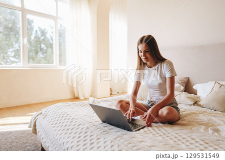 Young woman working on laptop while sitting on bed in bright, cozy bedroom during daytime 129531549