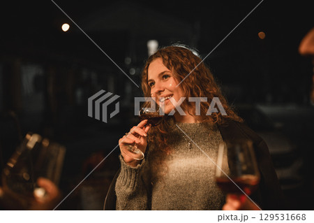 Woman smiling while holding a glass of wine at a nighttime gathering with friends outdoors 129531668