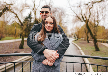 Couple embraces on a scenic bridge during a cloudy day in a tranquil park with trees in early spring 129531752