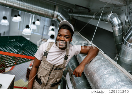 Friendly worker in casual clothing smiles while leaning against industrial piping indoors Friendly worker in casual clothing smiles while leaning against industrial piping indoors 129531840
