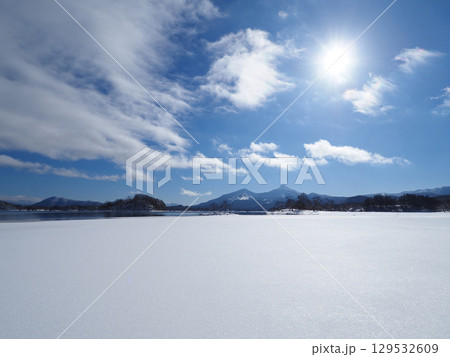 冬の裏磐梯、桧原湖。湖面は半凍結し、白い雪原が広がる。冠雪の磐梯山と青空に輝く太陽が印象的で綺麗だ。 129532609