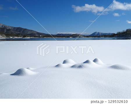 冬の裏磐梯、桧原湖。半分凍結した白い雪原に雪の小さな山できた。遠方には雪山と青い湖面が印象的で綺麗だ 129532610