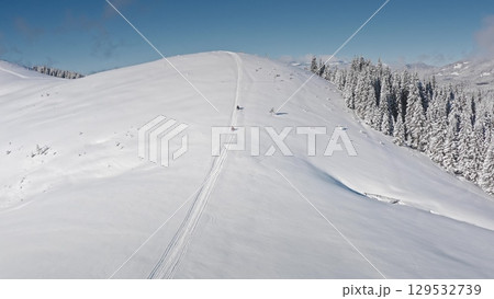 Two snowmobiles driving up a snowy mountain slope, leaving tracks in fresh snow, on a sunny day with clear blue sky and snowy forest on the side. Bukovel ski resort. Winter nature travel background 129532739