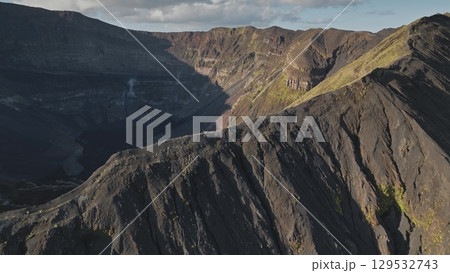 Drone captures rugged beauty of a volcanic crater, smoke and steam rising from its depths as clouds drift across sky, casting shadows on dramatic landscape. Amazing nature travel background Drone captures rugged beauty of a volcanic crater, smoke and steam rising from its depths as clouds drift across sky, casting shadows on dramatic landscape. Amazing nature travel background 129532743
