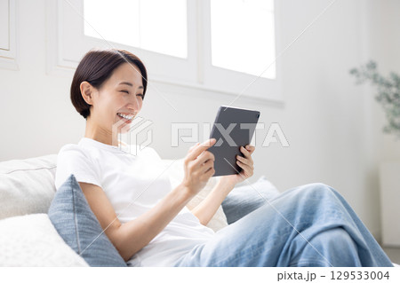 明るいリビングでソファに座りタブレットを操作し笑顔を見せる女性 129533004