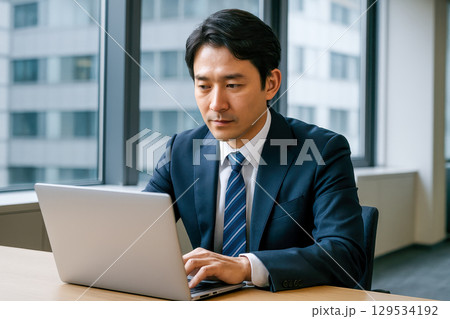 Focused asian businessman working on laptop in modern office, serious japanese male professional typing on computer, concentrated manager analyzing data on screen 129534192