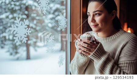 Woman enjoying hot chocolate in winter looking out window with snowflakes. Cozy hygge moment with female holding mug of cocoa on cold snowy day 129534193