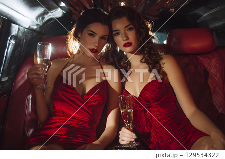 エレガントな赤いドレスを着た女性2人がリムジンでシャンパンを楽しむナイトシーン 129534322