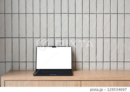 Laptop with a blank white screen placed on a wooden cabinet against a tile wall background 129534697