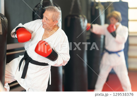 Elderly man in white kimono practices punching punching bag Elderly man in white kimono practices punching punching bag 129538376