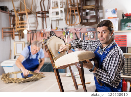 Middle-aged male restorer removes old cloth seat covering from antique wooden chair. 129538910