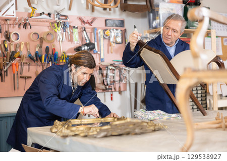 Two professional repairmen carpenters in uniform renewing chair mirror frame furniture in woodwork studio Two professional repairmen carpenters in uniform renewing chair mirror frame furniture in woodwork studio 129538927