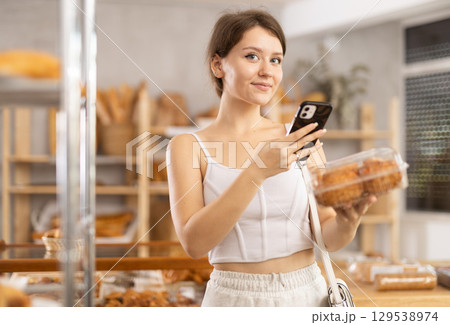 Cute young girl choosing pastries in bakery store, scans QR code on packaging box using mobile 129538974