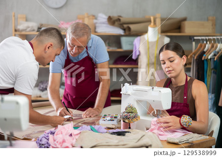Girl master sew clothes in sewing machine, senior craftsman with guy helper draw dress pattern Girl master sew clothes in sewing machine, senior craftsman with guy helper draw dress pattern 129538999