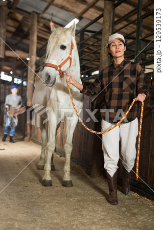 Asian woman leads a white horse by the bridle from stable 129539173