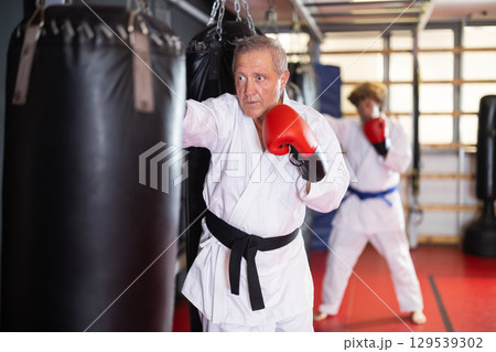 Elderly man in white kimono practices punching punching bag 129539302