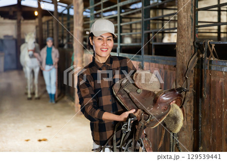 Asian female riding instructor walking in stable with saddle in hands Asian female riding instructor walking in stable with saddle in hands 129539441