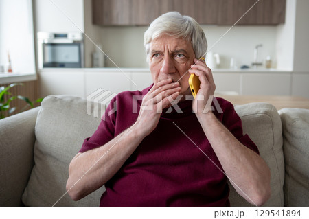 Suspicious elderly man with phone at home, targeted by scam artists, telemarketing fake news 129541894