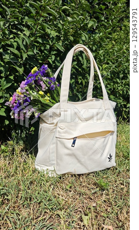 A white canvas tote bag with a bouquet of colorful wildflowers rests on green grass under bright sunlight. This image symbolizes eco-friendly lifestyle, freshness, and summer vibes 129543791