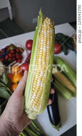 A fresh ear of bi-color corn is held in hand with husks partially peeled, surrounded by colorful vegetables and fruits 129543795