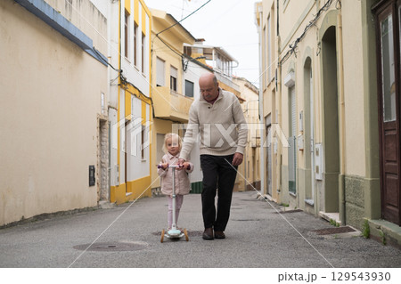 A grandfather is riding a scooter with his 3-year-old granddaughter on the street A grandfather is riding a scooter with his 3-year-old granddaughter on the street 129543930