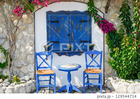 Mediterranean-style seating area features two blue wooden chairs, a small round table, and a rustic blue shuttered window 129544714