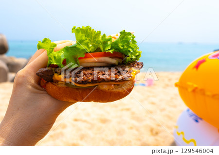 Woman holding burger in a hand against beach background 129546004