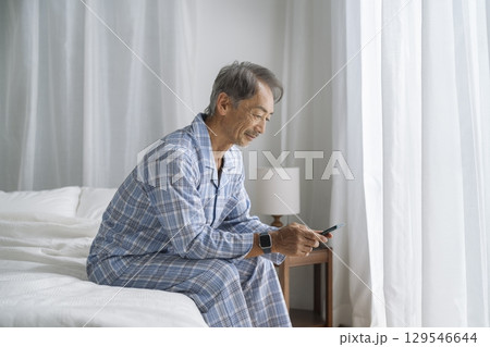 ベッドに座ってスマートフォンを操作するシニア男性 129546644