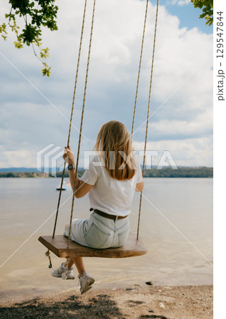 Blonde woman swinging gently on wooden swing above tranquil lake, savoring warmth of relaxing summer day while enjoying peaceful beauty of nature. Woman swinging on wooden swing, suspended over lake 129547844