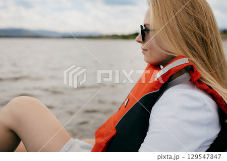 Young blonde woman wearing sunglasses and life jacket, enjoying scenic boat trip on tranquil lake while contemplating beautiful landscape and serene surroundings. Blonde woman relaxing in life jacket Young blonde woman wearing sunglasses and life jacket, enjoying scenic boat trip on tranquil lake while contemplating beautiful landscape and serene surroundings. Blonde woman relaxing in life jacket 129547847