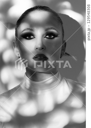 Black and white image of beautiful woman with fancy make-up standing in shadow and looking at camera 129548308