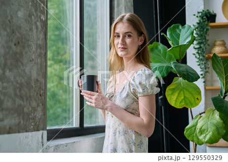 Young beautiful smiling romantic girl looking at camera with cup, standing near window Young beautiful smiling romantic girl looking at camera with cup, standing near window 129550329
