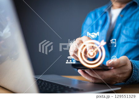 A businessman in a blue shirt interact with a smartphone, displaying holographic business icons, including a glowing target symbolizing goal achievement, analytics, partnerships, and financial growth. 129551329