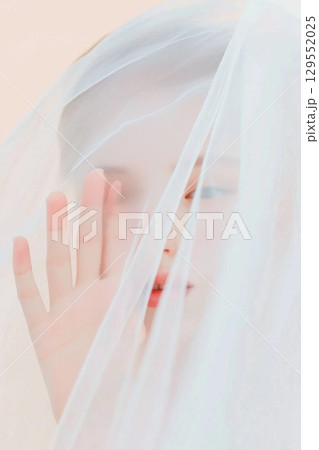 Close up portrait of a woman with short hair hidden behind a white veil, touching it with her hand, creating a sense of mystery and femininity 129552025