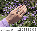 Landscape photo of woman's hand wearing a large oval cut diamond engagement ring, showcased against a blurred background of vibrant purple flowers in a sunlit garden. 129553308
