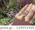 Close up landscape photo of woman's hand wearing a large oval cut diamond engagement ring, showcased against a blurred background of vibrant purple flowers in a sunlit garden. 129553309