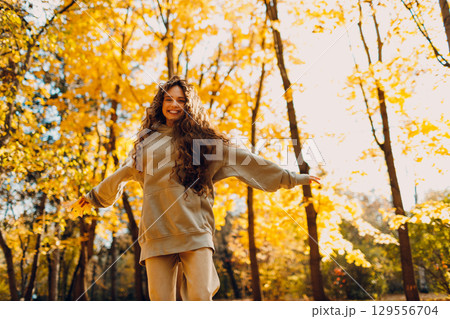 Smiling young woman plays and jumping dancing in the autumn forest with the yellow leaves at sunset Smiling young woman plays and jumping dancing in the autumn forest with the yellow leaves at sunset 129556704