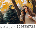 Smiling young woman photographer with the photo camera shooting the forest trees with the yellow leaves at sunset 129556728