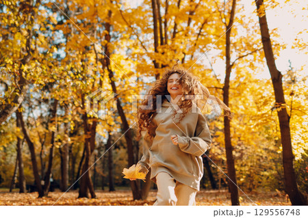 Smiling young woman plays and jumping dancing in the autumn park with the yellow leaves at sunset 129556748