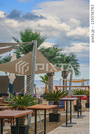 Outdoor Beach Cafe with Palm Trees, wooden tables, and decorative lights on sandy shore with cloudy sky. Concept of beach cafe, tropical seaside leisure, summer relaxation Outdoor Beach Cafe with Palm Trees, wooden tables, and decorative lights on sandy shore with cloudy sky. Concept of beach cafe, tropical seaside leisure, summer relaxation 129556763