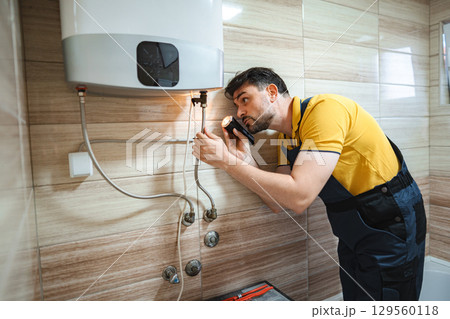 Plumber inspecting water heater with flashlight in bathroom 129560118