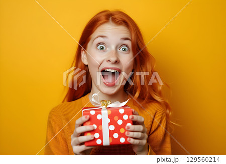 Excitement shines as a girl with red hair happily holds a gift on a vibrant yellow background 129560214