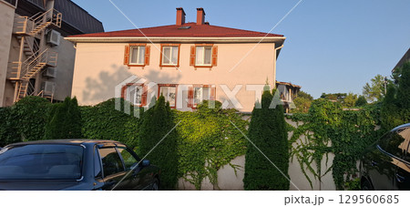 Modern house with a fence covered with vegetation.A three-story house with a red roof and a light facade, surrounded by a fence covered with climbing ivy. Modern house with a fence covered with vegetation.A three-story house with a red roof and a light facade, surrounded by a fence covered with climbing ivy. 129560685