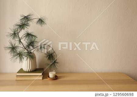 Minimalist Christmas interior still life. White vase with pine tree branches on stacked books, small candle in glass holder on wooden table against beige wall background. Warm winter home office. 129560698